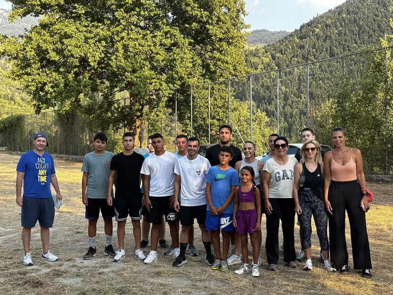 Ομαδική φωτογραφία των συμμετεχόντων στις αθλοπαιδιές στο γήπεδο του χωριού, με φόντο το βουνό